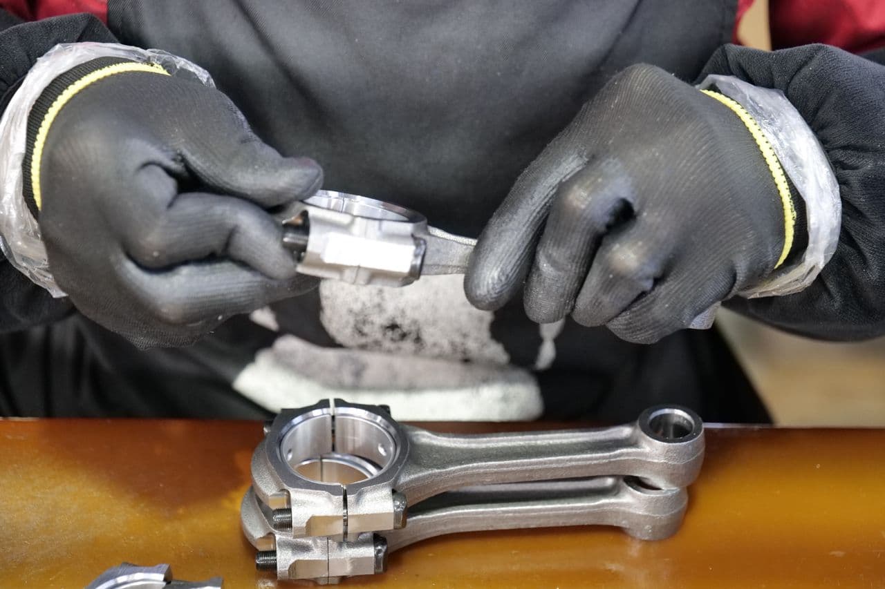 Gloved hands fitting a bearing cap to a machined connecting rod on a workbench.